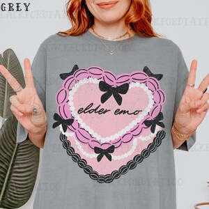 May include: Grey t-shirt featuring a pink heart-shaped graphic with black bows and the text "elder emo." The heart is outlined with a white beaded border and a black lace-like trim. The shirt has a relaxed fit.