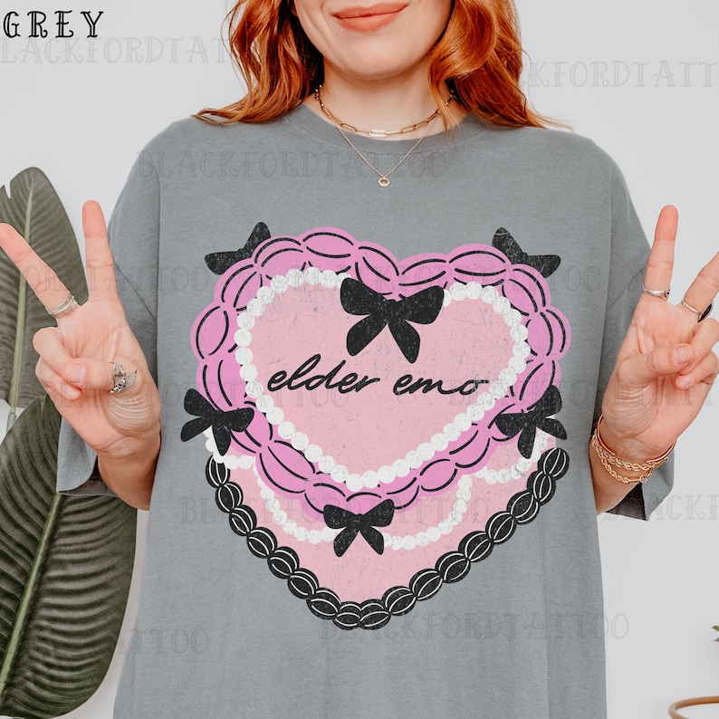 May include: Grey t-shirt featuring a pink heart-shaped graphic with black bows and the text "elder emo." The heart is outlined with a white beaded border and a black lace-like trim. The shirt has a relaxed fit.
