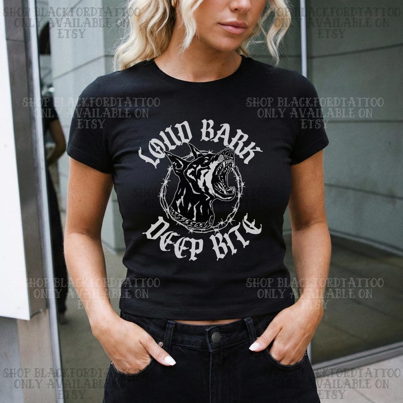 Puede incluir: Camiseta negra con un gr&aacute;fico blanco de un perro gru&ntilde;endo dentro de un c&iacute;rculo de alambre de p&uacute;as. El texto "LOUD BARK DEEP BITE" est&aacute; encima y debajo de la ilustraci&oacute;n del perro. La camiseta es de manga corta.