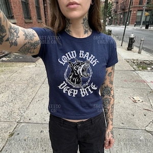 Puede incluir: Camiseta azul marino con el texto "LOUD BARK DEEP BITE" y un gr&aacute;fico de un perro gru&ntilde;endo dentro de un c&iacute;rculo de alambre de p&uacute;as. La camiseta es de estilo ajustado.