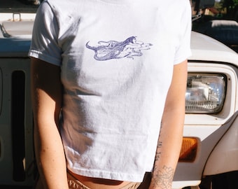 May include: White crew neck t-shirt featuring a purple illustration of a mermaid. The graphic shows a mermaid with long flowing hair and a fishtail, swimming in the water. The t-shirt is made of a soft, lightweight material.
