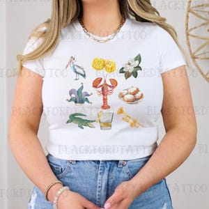 May include: White t-shirt with a colorful illustration of various symbols associated with Mardi Gras, including a pelican, a fleur-de-lis, a crawfish, a glass of Hurricanes, and beignets.