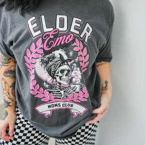 May include: A gray t-shirt with a pink graphic of a skeleton with a bun in their hair wearing a bandana and holding a cup of coffee. The graphic also includes the text "Elder Emo Moms Club".