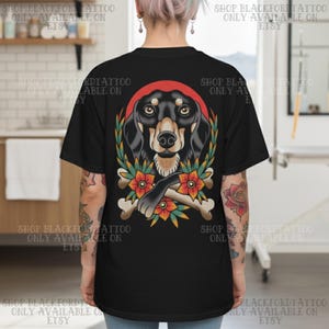 K&ouml;nnte beinhalten: Schwarzes T-Shirt mit einer Tattoo-Illustration eines Dackelhundes. Das Design umfasst einen roten Heiligenschein, florale Elemente und gekreuzte Knochen. Der Text "SHOP BLACKFORDTATTOO ONLY AVAILABLE ON ETSY" ist um das Shirt gedruckt.