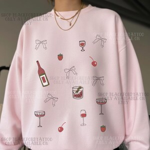 Op de afbeelding: Zachtroze sweatshirt met een herhaald patroon van wijnglazen, strikken, kersen, aardbeien en een wijnfles. De tekst "SHOP BLACKFORDTATTOO ONLY AVAILABLE ON ETSY" is rondom gedrukt.
