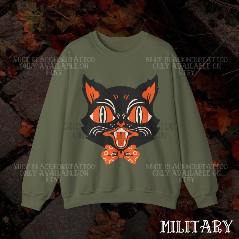 May include: Olive green sweatshirt featuring a black cat face with orange eyes, ears, and a bow tie. The cat has an open mouth with visible teeth. Text on the sweatshirt reads "SHOP BLACKFORDTATTOO ONLY AVAILABLE ON ETSY" and "MILITARY".