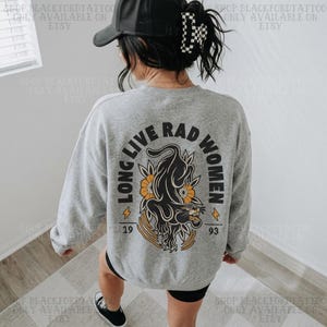 May include: A heather grey long-sleeve sweatshirt featuring a black panther graphic with orange floral accents and the text LONG LIVE RAD WOMEN 19 93. The garment is worn with a black baseball cap that has a white and black chequered pattern on the side.