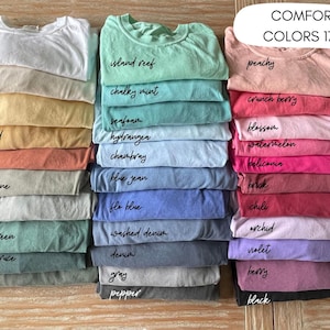 May include: A stack of folded t-shirts in various colors, including white, mustard, and blue. Each shirt is labeled with its color name in a handwritten font. The text "COMFORT COLORS 1717" is displayed at the top right.