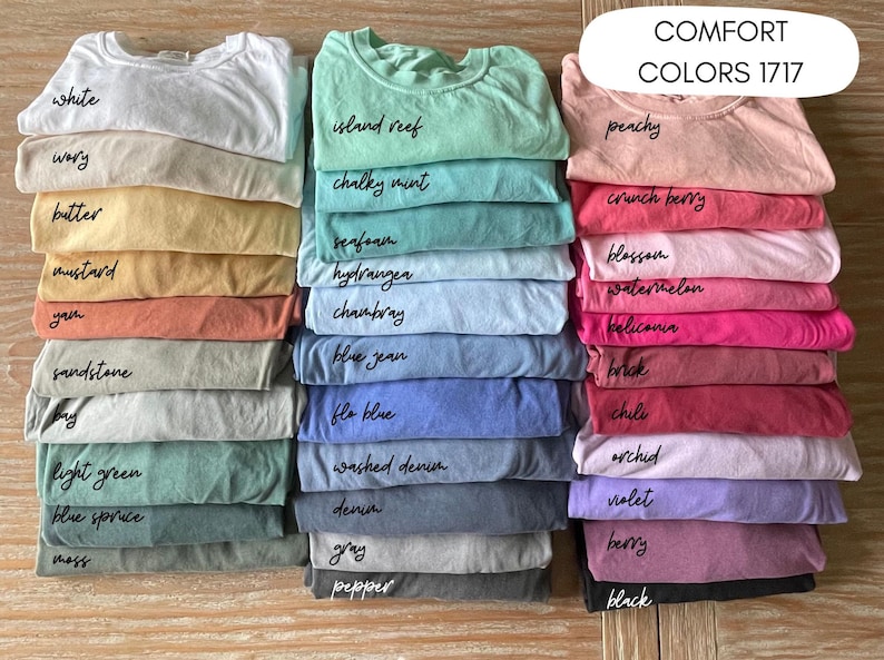 May include: A stack of folded t-shirts in various colors, including white, mustard, and blue. Each shirt is labeled with its color name in a handwritten font. The text "COMFORT COLORS 1717" is displayed at the top right.