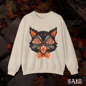 May include: Sand-colored sweatshirt featuring a black cat graphic with orange eyes, ears, and a bow tie. The cat has an open mouth with visible teeth. The text "SAND" is at the bottom. The sweatshirt has long sleeves.