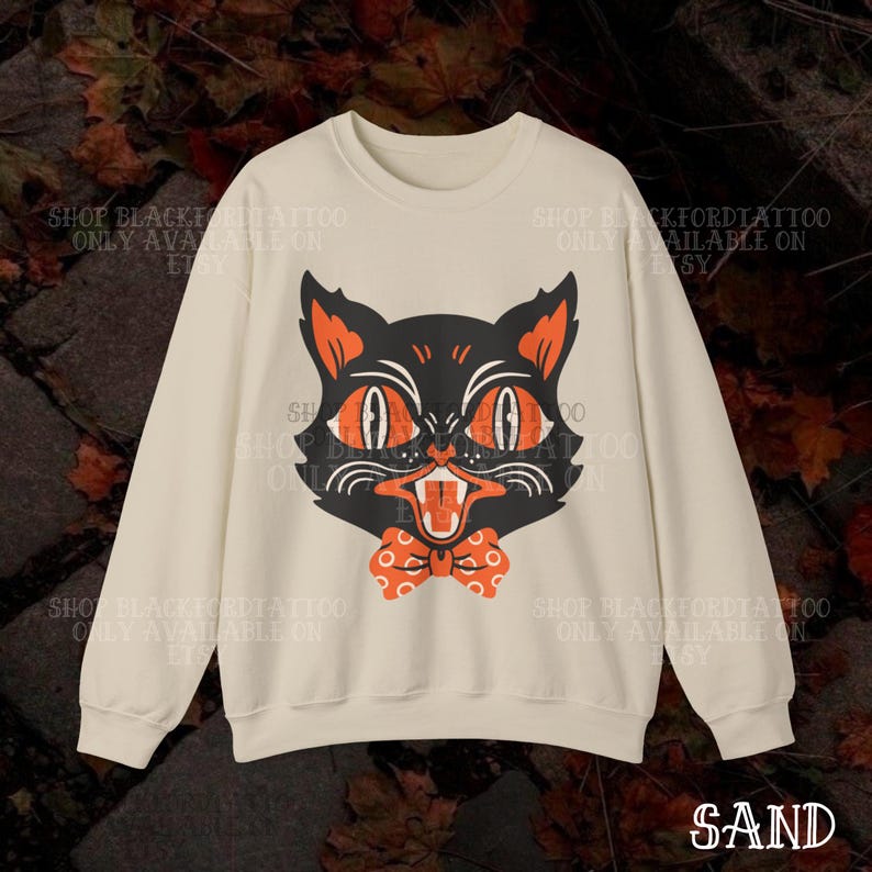 May include: Sand-colored sweatshirt featuring a black cat graphic with orange eyes, ears, and a bow tie. The cat has an open mouth with visible teeth. The text "SAND" is at the bottom. The sweatshirt has long sleeves.