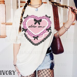 May include: Ivory-colored t-shirt featuring a pink heart design with black bows and the text "elder emo." The heart is outlined with a black scalloped border and white pearls. The shirt has short sleeves.
