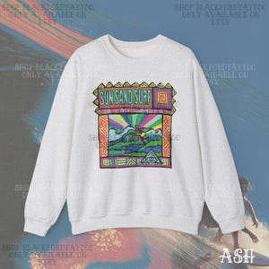 May include: A light gray sweatshirt with a colorful graphic that says "SUN SAND SURF". The graphic features a surfer, waves, and a rainbow. The sweatshirt has long sleeves and a crew neck.
