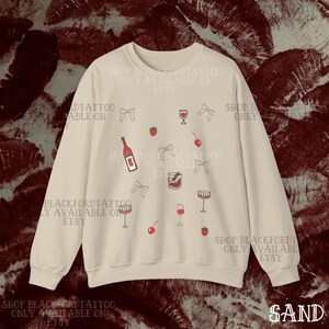 May include: Sand-colored sweatshirt with a repeating pattern of wine bottles, glasses, cherries, strawberries, and bows. The text "SHOP BLACKFORDTATTOO ONLY AVAILABLE ON ETSY" is printed around the design. The word "SAND" is printed at the bottom right.