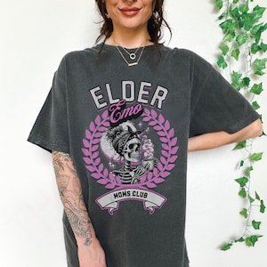 May include: A dark grey t-shirt with a pink graphic design featuring a skull with a bandana and a coffee cup. The text "Elder Emo Moms Club" is printed on the shirt.