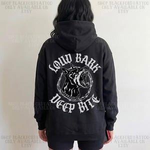 Puede incluir: Sudadera con capucha negra con un gráfico blanco de una cabeza de perro gruñendo rodeada de alambre de púas, y las palabras "LOUD BARK DEEP BITE". Prenda de vestir informal.