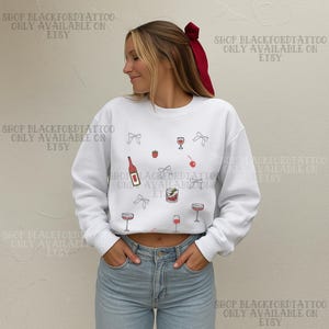May include: White sweatshirt with a cocktail-themed design. The design includes illustrations of wine glasses, a bottle, cherries, strawberries, and bows. The text "SHOP BLACKFORDTATTOO ONLY AVAILABLE ON ETSY" is printed on the top and bottom of the image.