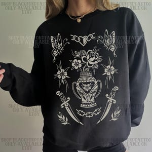 Może przedstawiać: Czarna bluza z białym wzorem w stylu tatuażu. Wzór zawiera wazon z kwiatami, motyle, sztylety i elementy kwiatowe. Tekst "SHOP BLACKFORDTATTOO ONLY AVAILABLE ON ETSY" jest wydrukowany.
