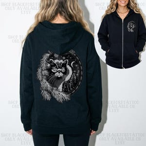 Puede incluir: Una sudadera con capucha y cremallera en azul marino oscuro que presenta un gran gráfico circular en la espalda con una cara de gato, ramas de pino y una luna creciente. Una versión más pequeña del mismo gráfico aparece en el pecho izquierdo.