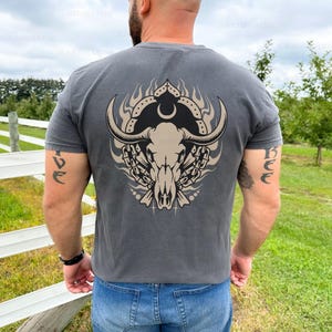Puede incluir: Una camiseta gris antracita con un gr&aacute;fico de un cr&aacute;neo de toro con cuernos, llamas y una luna creciente en la espalda.