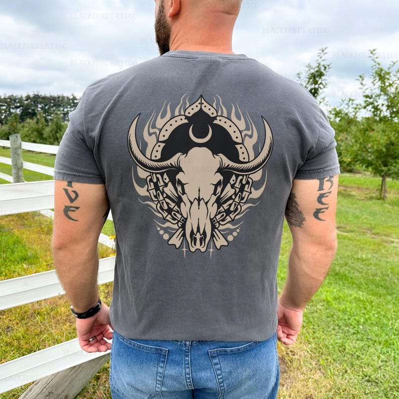Puede incluir: Una camiseta gris antracita con un gr&aacute;fico de un cr&aacute;neo de toro con cuernos, llamas y una luna creciente en la espalda.