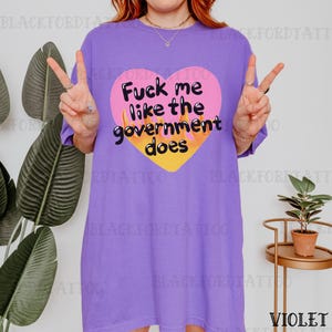 Puede incluir: Camiseta violeta con la frase "Fuck me like the government does" dentro de un corazón rosa con llamas amarillas. La camiseta es de color liso y tiene mangas cortas.