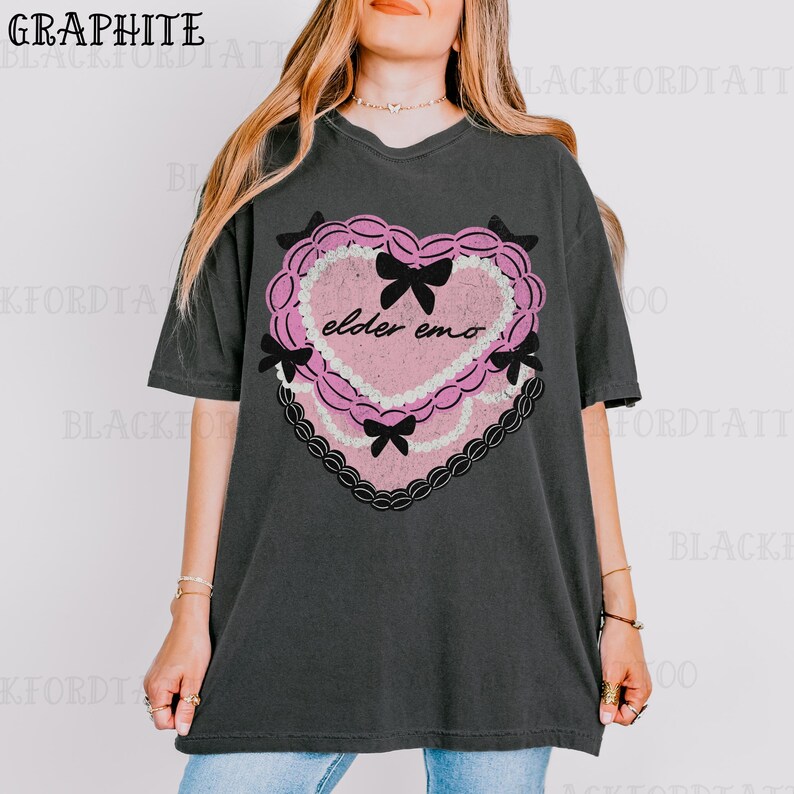May include: Graphite-colored t-shirt featuring a pink heart design with black bows and the text "elder emo." The heart has a lace-like border and a distressed texture. The shirt is a relaxed fit.