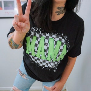 May include: Black t-shirt with a green "MAMA" graphic on a checkered background. The shirt has a distressed look with white paint splatters.