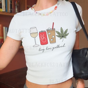 May include: White crop top with a graphic of a wine glass, soda can, iced coffee, and cannabis leaf. The text "long live girlhood" is below the graphic. The shirt has short sleeves.