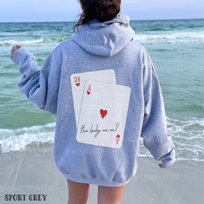 Puede incluir: Una sudadera gris con un gr&aacute;fico de dos cartas de juego en la espalda. Las cartas son un 28 y un As de Corazones. El texto en las cartas dice "How lucky are we?"
