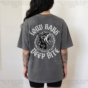Könnte beinhalten: Graues T-Shirt mit einem schwarzen und weißen Bild eines knurrenden Hundekopfes, umgeben von Stacheldraht. Der Text "LOUD BARK DEEP BITE" ist darüber und darunter gedruckt. Das T-Shirt hat eine lockere Passform.