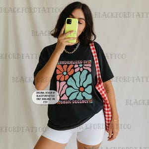 Può includere: T-shirt nero con grafica floreale arancione, rosa e turchese, con la scritta "RADICALIZED BY BASIC DECENCY". La maglietta è abbinata a pantaloncini bianchi e una borsa tote a scacchi rossi e bianchi. Una persona tiene in mano un telefono giallo.