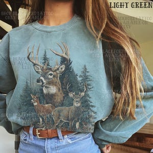 May include: A light green crewneck sweatshirt featuring a graphic print of three deer standing in a forest of pine trees. The design includes a large buck with antlers, a smaller deer, and a fawn, all rendered in natural brown tones against the fabric.