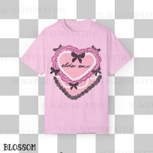 May include: A light pink t-shirt featuring a heart-shaped design with the words "elder emo" in the center. The heart is decorated with black bows, a pearl-like border, and a black lace trim. The word "BLOSSOM" is at the bottom.
