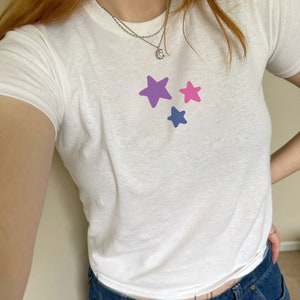May include: White t-shirt with three colorful star graphics in purple, pink, and blue. The shirt is paired with blue denim jeans. A silver necklace is worn around the neck.