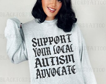 May include: A pale grey sweatshirt featuring the text "SUPPORT YOUR LOCAL AUTISM ADVOCATE" in a gothic font. The word "ASH" is printed at the bottom left corner. The sweatshirt is long-sleeved with a crew neck.