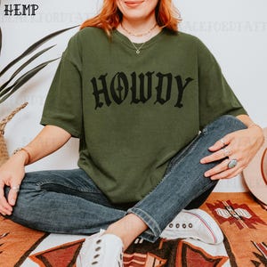 May include: A woman wearing a green t-shirt with the word "Howdy" printed in black gothic lettering. She is sitting on a colorful rug with her legs crossed and wearing blue jeans and white sneakers.