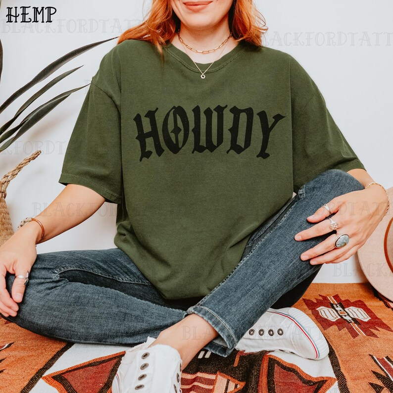 May include: A woman wearing a green t-shirt with the word "Howdy" printed in black gothic lettering. She is sitting on a colorful rug with her legs crossed and wearing blue jeans and white sneakers.