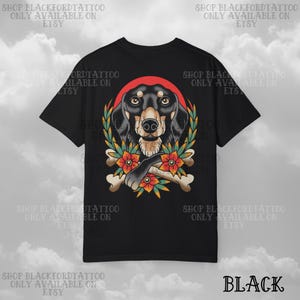 K&ouml;nnte beinhalten: Schwarzes T-Shirt mit einer detaillierten Illustration eines Dackelhundes, umgeben von einem roten Heiligenschein, einem Lorbeerkranz und Blumen. Die Pfote des Hundes liegt auf gekreuzten Knochen. Der Text "BLACK" steht unten.