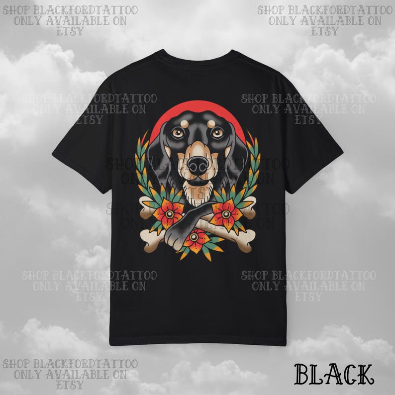 K&ouml;nnte beinhalten: Schwarzes T-Shirt mit einer detaillierten Illustration eines Dackelhundes, umgeben von einem roten Heiligenschein, einem Lorbeerkranz und Blumen. Die Pfote des Hundes liegt auf gekreuzten Knochen. Der Text "BLACK" steht unten.