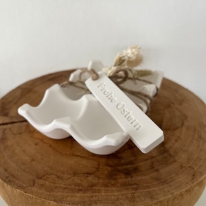 Könnte beinhalten: Ein weißer Keramik-Eierhalter mit zwei Fächern, verziert mit einem Anhänger mit der Aufschrift "Frohe Ostern". Der Halter ist mit Bindfaden und getrocknetem Weizen gebunden. Er steht auf einem runden Holzsockel.