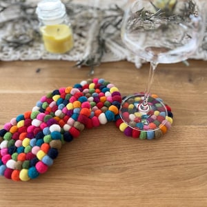 Könnte beinhalten: Ein farbenfrohes Filzkugel-Untersetzer-Set mit einem länglichen und einem runden Untersetzer. Die Untersetzer bestehen aus kleinen, bunten Filzkugeln in verschiedenen Farben, darunter Rot, Blau, Grün und Gelb. Ein Weinglas steht auf dem runden Untersetzer.