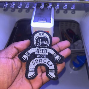 May include: Black and white embroidered patch in the shape of an astronaut. The patch has the words "You NEED more SPACE?" in a circular design. The patch is being held in a hand.