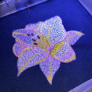 May include: Close-up of an embroidered flower in shades of purple, yellow, and pink. The floral design is on a dark blue fabric, with the embroidery machine's needle and frame visible in the background. The flower has detailed stitching.