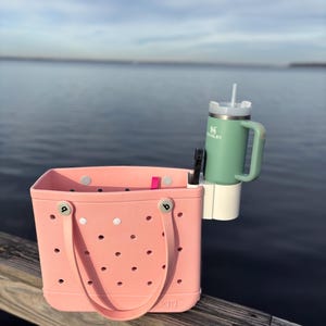 Op de afbeelding: Een lichtroze draagtas met een handvat, een groene Stanley-mok met een wit rietje en een witte bekerhouder. De draagtas heeft verschillende kleine gaatjes en staat op een houten oppervlak.