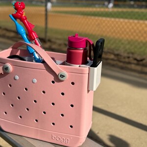 Op de afbeelding: Een roze draagtas met een geperforeerd ontwerp, met een rode waterfles, een zwarte telefoonhouder en twee bellenblaasstokken. De tas staat op een metalen bank in de buurt van een honkbalveld.