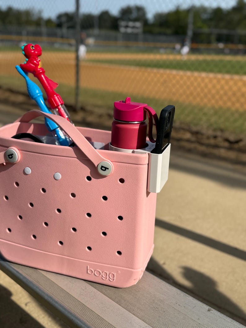 Op de afbeelding: Een roze draagtas met een geperforeerd ontwerp, met een rode waterfles, een zwarte telefoonhouder en twee bellenblaasstokken. De tas staat op een metalen bank in de buurt van een honkbalveld.