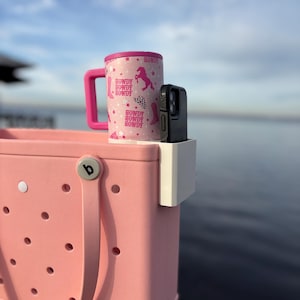 Op de afbeelding: Een roze draagtas met een witte telefoonhouder en een roze en witte reismok. De mok heeft een roze deksel en handvat, met een herhalend patroon van cowboylaarzen, paarden en het woord "Howdy". De telefoonhoes is zwart.