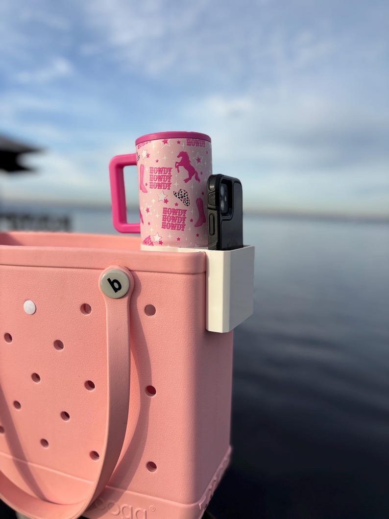 Op de afbeelding: Een roze draagtas met een witte telefoonhouder en een roze en witte reismok. De mok heeft een roze deksel en handvat, met een herhalend patroon van cowboylaarzen, paarden en het woord "Howdy". De telefoonhoes is zwart.