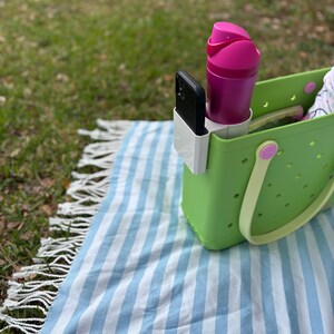 Op de afbeelding: Een limoengroene draagtas met een witte telefoonhouder, een magenta waterfles en een zwarte smartphone. De tas staat op een blauw-wit gestreepte deken met een gefranjerde rand, op een grasachtig oppervlak.